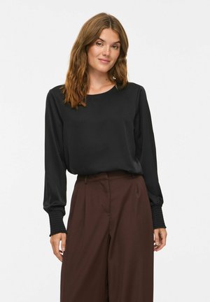 Donna con capelli castani ondulati che indossa una camicetta nera a maniche lunghe con polsini arricciati e pantaloni marroni a gamba larga e vita alta.