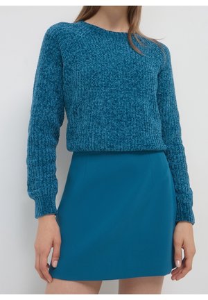Femme portant un pull bleu texturé à manches longues et une jupe bleue ajustée unie sur un fond clair uni.