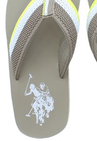 Tan flip-flopit, jossa on verkkoansat ja valkoisia sekä keltaisia yksityiskohtia. Sisältää grafiikan kahdesta polo-pelaajasta hevosilla. Tasapohjainen muotoilu.