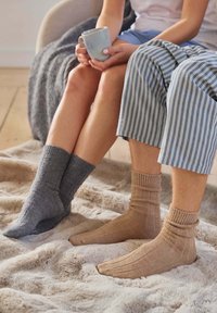 Šedé a béžové pletené ponožky s žebrovanými manžetami a měkkou texturou, oblečené s pruhovanými pyžamovými kalhotami, odpočívající na načechrané dece.