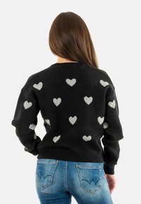 Maglione lavorato a maglia nero con motivi a cuore argento, colletto rotondo, spalle basse e polsini a costine. Indossato con jeans blu con cuciture decorative.