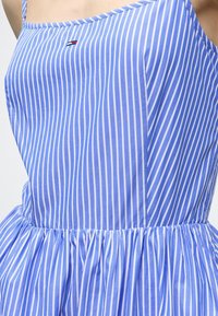 Robe sans manches à rayures verticales bleues et blanches, avec un corsage ajusté et une taille froncée, montrée de la moitié de la poitrine à la taille.