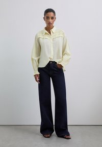 Chemise jaune clair avec fermeture à boutons, devant texturé et manches bouffantes, assortie à un jean en denim foncé à jambe large. Le modèle se tient sur un fond neutre.
