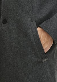 Abrigo de lana gris oscuro con un solo botón, bolsillo lateral y acento metálico con el nombre de la marca. Tejido texturizado, ajuste entallado.