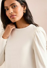 Krémová blůza s kulatým výstřihem, dlouhými nabíranými rukávy, jemnou texturou a minimalistickým designem. Modelka si upravuje vlasy s přirozeným make-upem.