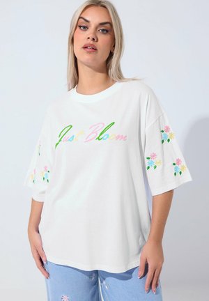 Lyshåret kvinde iført en hvid oversized T-shirt med teksten "Just Bloom" i pastelfarver og blomstret broderi på ærmerne samt lyseblå jeans.