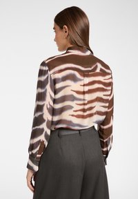 La blusa di seta presenta un motivo marrone e crema con strisce astratte. Maniche lunghe con polsini bottonati e dettaglio di piega sul retro.