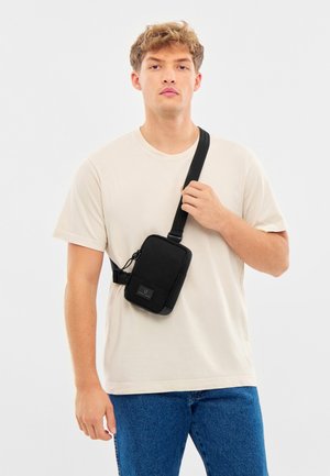 Jonge man draagt een beige t-shirt en blauwe spijkerbroek met een zwarte crossbodytas, hij houdt het bandje vast met één hand.
