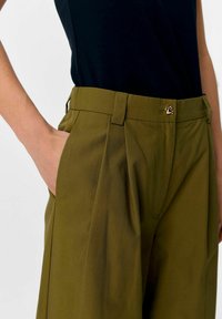 Olivgrüne Hose mit vorderen Falten, flachem Bund und einem metallischen Verschluss an der Taille. Hergestellt aus einem glatten, leichten Stoff.