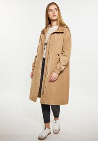 Beige vattentålig regnjacka med huva, framknappar, elastiska ärmslut och fickor, bärs över en randig topp och mörka byxor. Vita sneakers.