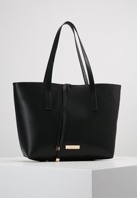 Sac fourre-tout en cuir noir avec double poignée, texture lisse, fermeture éclair sécurisée et une petite plaque dorée gravée "ANNA FIELD".