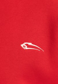 Červená tkanina s hladkým povrchem obsahuje malé bílé logo vyšitěné na levé straně, které ukazuje elegantní, stylizovaný design.