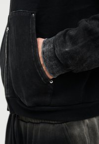 Μαύρο ζακέτα με φερμουάρ, με μανσέτα από υφασμάτινη υφή σε γκρι χρώμα, πλαϊνή τσέπη και μεταλλικό κουμπί κοντά στη ραφή της τσέπης.