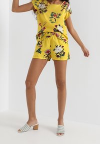 Ensemble haut et short imprimés à fleurs jaunes, en tissu lisse. Sandales blanches à enfiler avec accents texturés et petit talon carré.