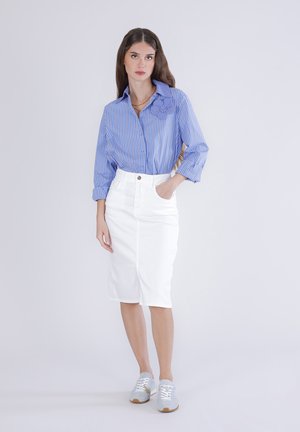 Kvinde med langt brunt hår iført en blåstribet skjorte, hvid knælængde nederdel og grå sneakers, stående med den ene hånd i lommen.