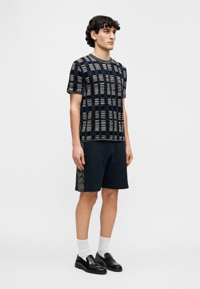 Μπλε πουκάμισο με σχέδιο και κοντά μανίκια, με υφή πλέγματος, συνδυασμένο με μπλε σορτς με πλευρική λεπτομέρεια. Μαύρα μοκασίνια και λευκές κάλτσες.