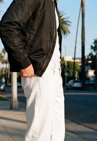 Personne portant une veste noire et un pantalon blanc, une main dans la poche, debout à l'extérieur près d'un trottoir avec des palmiers en arrière-plan.