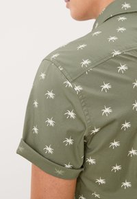 Camisa de hombre de manga corta de color verde, hecha de algodón, con un diseño estampado que presenta pequeñas palmeras blancas, mangas enrolladas y un corte relajado.