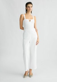 Une combinaison blanche à jambes larges, décolleté carré et détail de ceinture tressée. Modèle portant des chaussures à motifs, debout sur un fond neutre.