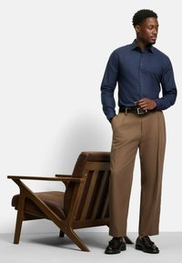 Blaues Hemd mit Knopf, khakifarbene Hose, schwarze Dressschuhe. Holzgestellstuhl mit brauner Polsterung. Kontrast zwischen Stoff und Farben.