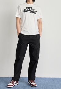 Vit bomullströja med svart "JUST DO IT"-grafik, kombinerad med lösa svarta cargobyxor och röda och vita träningsskor.