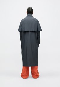 Dlhý sivý kabát s vrstveným dizajnom, vybavený zadným pelerínovým dielom. Kombinovaný s voľnými, jasne oranžovými nohavicami. Hladká textúra, minimalistický štýl.
