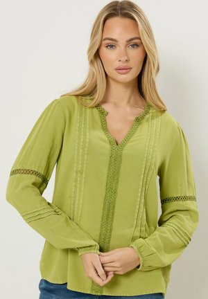 Jonge vrouw met lang blond haar, gekleed in een lichtgroene blouse met lange mouwen en kantdetails, en een blauwe spijkerbroek, handen in elkaar gevouwen ter hoogte van de taille.