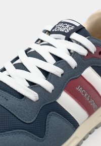 Blå sneaker med mesh- og ruskindstekstur, hvide snørebånd, røde og hvide sidelæstreger samt et mærket label på tungen. Retro design.