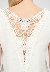Hvit topp med V-formet blonderygg med blomstermotiver og intrikate mønstre. Myk tekstur med et avslappet design.