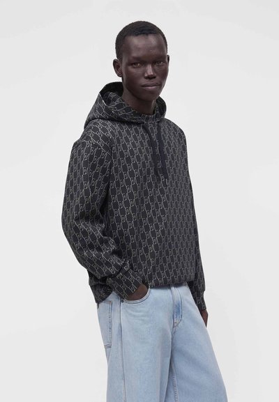 Jeune homme portant un sweat à capuche noir à motifs et un jean bleu clair, debout avec une main dans la poche devant un fond clair uni.