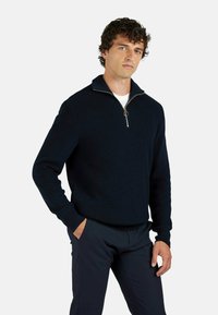Maglione blu navy a maglia con colletto a mezza zip, tessuto texturizzato e tirazip in metallo. Indossato con pantaloni scuri, caratterizzati da una silhouette aderente.