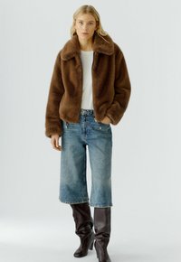 Blonde vrouw in een bruine nep-bont jas, witte top, knie-lange denim shorts en donkerbruine hoge laarzen op een effen achtergrond.