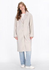 Cappotto trench chiaro beige con colletto a scollo e due bottoni, dotato di due tasche laterali, indossato sopra una maglietta bianca e jeans blu.