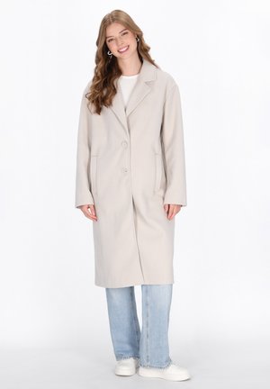 Cappotto trench chiaro beige con colletto a scollo e due bottoni, dotato di due tasche laterali, indossato sopra una maglietta bianca e jeans blu.