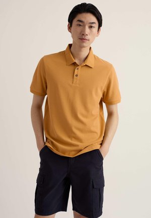 Noor mees seisab käed taskutes, kandes sinepikollast polo särki ja musti cargo lühikesi pükse, tavalise beeži tausta ees.