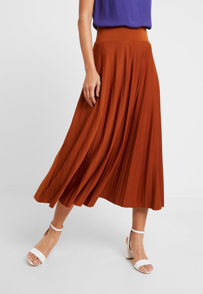 Femme portant une jupe midi plissée couleur rouille, un haut violet et des sandales blanches à talons blocs avec bride à la cheville, debout devant un fond uni.