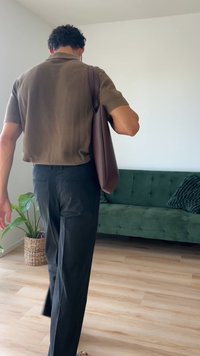 Ruda odinė petinė rankinė su apvalia forma, derinama su tamsiomis kelnėmis ir trumpomis rudomis marškinėliais, šviesiame kambaryje su žalia sofa.