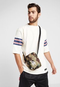 Borsa a tracolla camouflage verde e marrone con logo bianco, tracolla regolabile, chiusura a zip e piccola toppa. Indossata su una maglietta bianca a maniche corte.