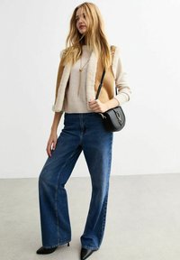 Tan shearlingväst över en ljusbeige stickad tröja, i kombination med högmidjade blå vida jeans och svarta spetsiga klackskor. Svart axelväska.