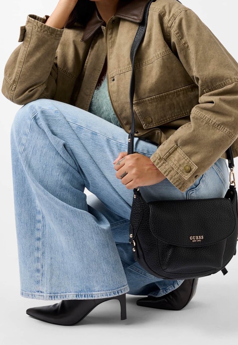Personne portant un jean bleu clair à jambes larges, une veste marron, des bottes noires à talons et tenant un sac à bandoulière noir Guess.