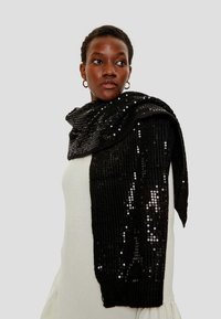 Sciarpa nera in lurex drappeggiata su un maglione chiaro a costine. Presenta una texture lucida con paillettes scintillanti e una forma allungata.