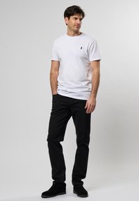 Cămașă albă din bumbac cu un mic logo bleumarin, asortată cu blugi negri din denim. Modelul poartă pantofi negri din suede, stând în fața unui fundal deschis.
