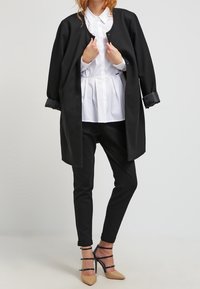 Manteau noir oversize avec manches retroussées, chemise blanche boutonnée, et pantalon noir. Le mannequin porte des talons beiges avec des accents de bride noire.