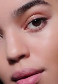 Närbild av ett ansikte med slät hy, en ljusrosa läppfärg och naturlig ögonmakeup, med täta mörka ögonbryn och en mjuk, neutral bakgrund.