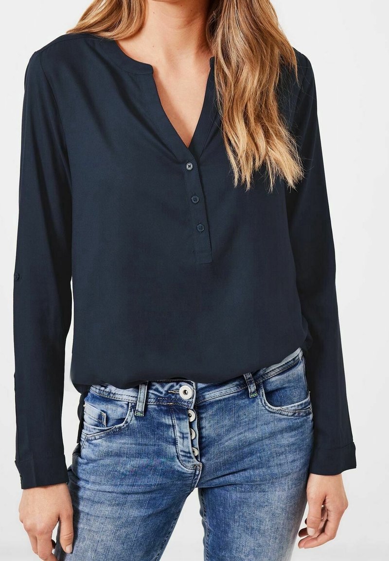 Femme portant un t-shirt henley bleu marine à manches longues avec trois boutons et un jean en denim bleu avec une braguette à boutons apparents.