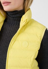 Chaleco acolchado amarillo con un cuello alto, con un suéter de cuello alto gris texturizado debajo y un parche con el logo en el pecho.