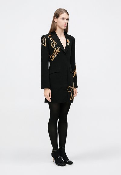 Rochie sacou neagră cu expresii din paiete aurii, închidere cu un singur nasture, două buzunare, mâneci lungi, asortată cu dresuri negre opace și pantofi cu toc.