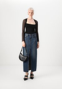 Schwarzes Langarmoberteil, hoch taillierter Jeansrock mit Schlitz vorne, schwarze gemusterte Handtasche und spitze schwarze Absatzschuhe mit Schnallen.