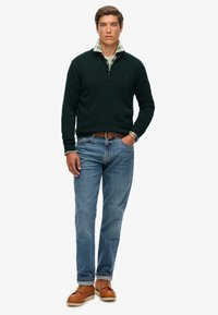 Maglione zip-up verde scuro sopra una camicia a scacchi con colletto, abbinato a jeans azzurri chiari e scarpe di pelle marrone. Vestibilità casual con toni neutri.