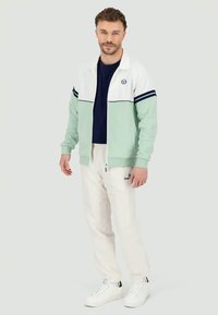 Giacca zippata verde menta e bianca con righe navy, abbinata a pantaloni jogger crema. Il modello indossa sneakers bianche e una maglietta blu scuro sotto.